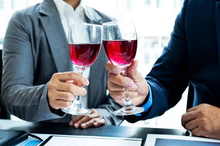 Two business executives people, businesswomen, and businessman in a suit is bumping into a glass of wine celebrating success at the office desk. After the meeting.の写真素材
