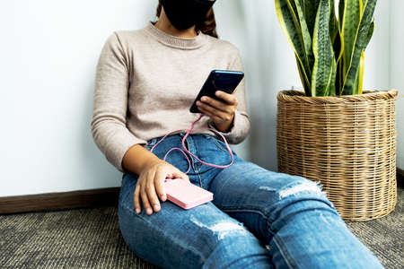young woman sitting against a wall holding a smartphone while charging a smartphone battery.の写真素材