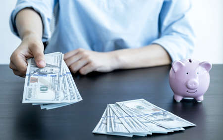The woman's hand holding the dollar note and there is a piggy bank on the table, step up start up business to success, Saving money for future plan and retirement fund conceptの写真素材