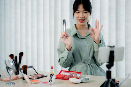 Beautiful Asia female vlogger is showing cosmetics makeup products while recording video and giving recommended for her beauty blog with digital smartphone.の写真素材