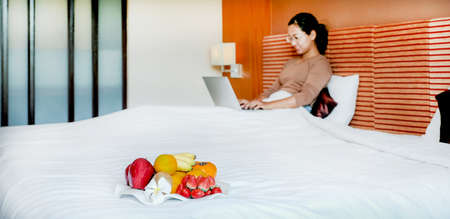 Fruits in tray in front of tourists used laptop on the bed in the luxury hotel room.