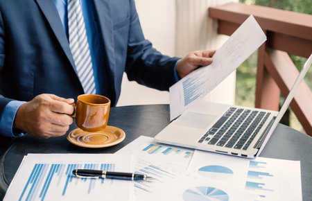 Businessman hand holding coffee cup, analysis the graph with laptop at the home office for setting challenging business goals And planning to achieve the new targetの写真素材