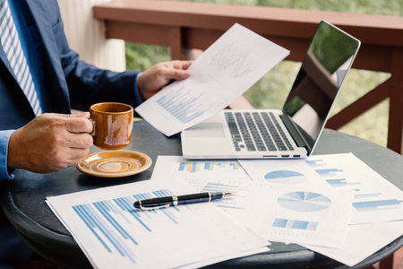 Businessman hand holding coffee cup, analysis the graph with laptop at the home office for setting...