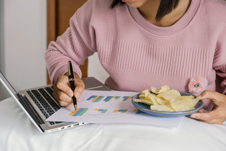 young woman hand holding pen used laptop video conferences and eating snack on the bed, healthy food and work from home concept.の写真素材