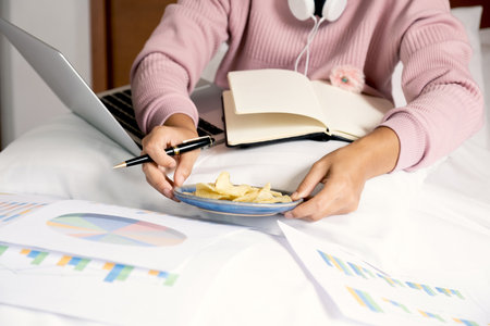 young woman hand holding pen used laptop video conferences and eating snack on the bed, healthy food and work from home concept.の写真素材
