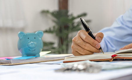 Businessman hand writing income and expenses to account with piggy bank and dollar notes, step up start-up businesses to success, Saving money for future plan and retirement fund concept.の写真素材