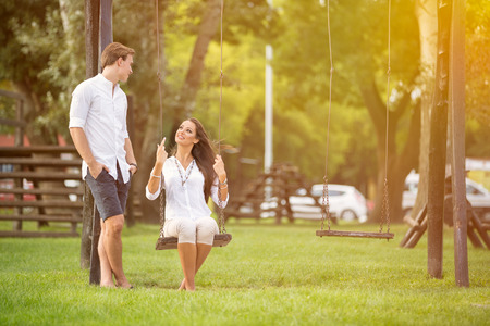 Attractive couple in park on swing, girl sitting on swing man standing next herの写真素材