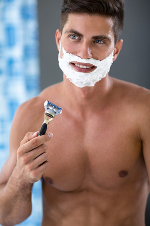 Young fit man with razor  front of mirror and shave foam on faceの写真素材