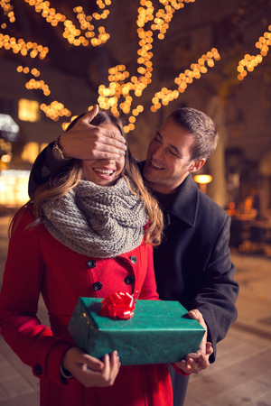 relationships, love, people, birthday and holidays concept - happy man covering womanâs eyes and giving gift box on streetの写真素材