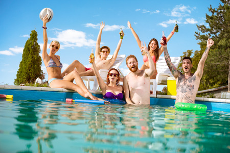 Cheerful young people in pool rejoice with beverage in raising hands in pool at summer