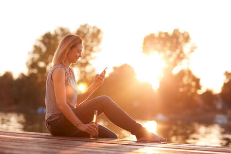 Alone lady sitting on dock near water with bottle and cellphone at sunsetの写真素材