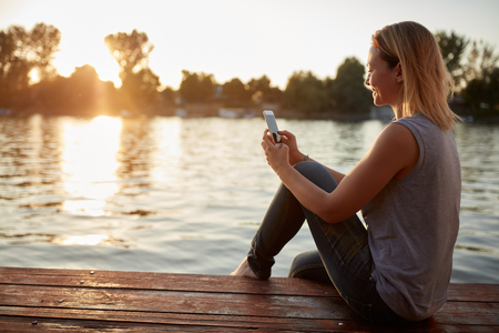 Woman with cellphone by river on sunsetの写真素材