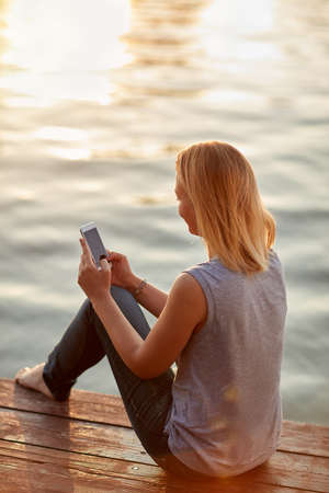 Young lady sitting on dock and looking in phoneの写真素材