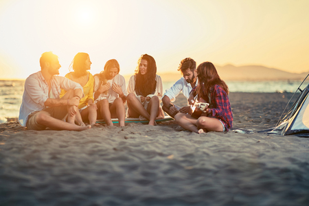 Group of young friends with guitar at beach. friends relaxing on sand at beach with guitar and singing on summer sunsetの写真素材
