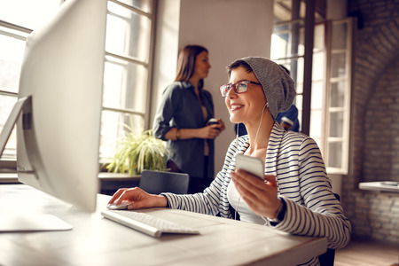 Young smile girl working on computer and listening music on cell phone in officeの写真素材