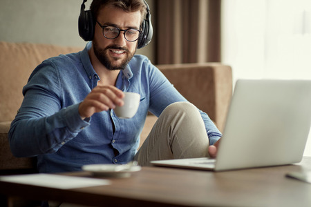 Young man with headphones using laptop and drinking coffee in his homeの写真素材
