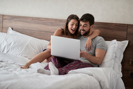 Happy young couple lying on a bed with computer - Beautiful married couple watching sex video on laptop laughing together - People, sexual, technology conceptの写真素材