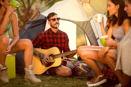 Hipster man on camping playing guitar in the company of his friendsの写真素材