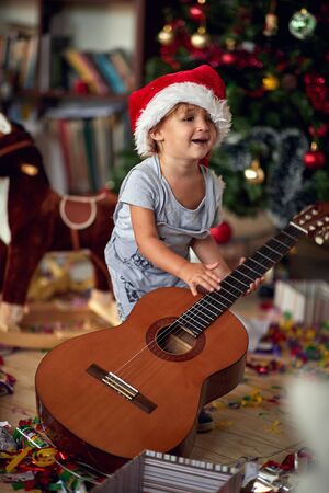 Cute boy in Santa hat is playing in front of a decorated Christmas tree with guitarの写真素材