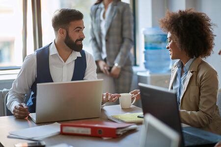 Colleagues exchanging ideas, talking, at the desk with their laptops in bright open space office.の写真素材