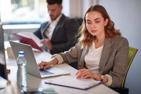 Businesswoman looking at the clipboard in the office. Woman working on a computer with her colleague in the backround. Data check.の写真素材