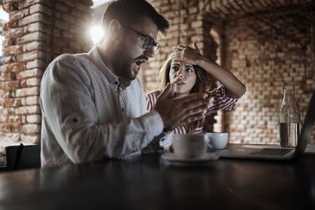 young caucasian couple looking at laptop in front of them, receiving bad news, made mistake, drinking coffee in cafeの写真素材