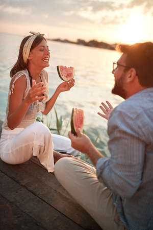 young couple sitting by the water talking, smiling, laughing, eating watermelon