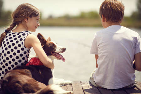 Little boy and girl with dog sitting on a wood pier and fishing in a pond.の写真素材