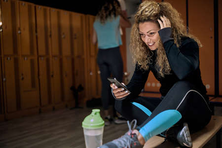 Smiling girl with a mobile phone sits  on the bench in the locker-room in the gym.の写真素材