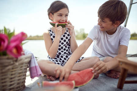 Cute kids  eating fresh watermelon on the pier.  Girl and boy enjoying together at lake.の写真素材