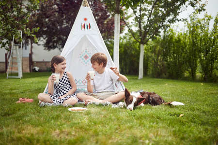 Cute happy little children sitting in teepee at backyard and drink milk.