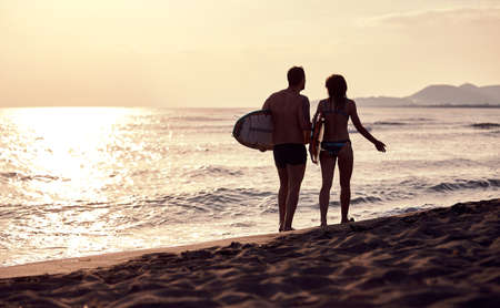 young caucasian couple walking on the sandy beach, carrying surfboards, talkingの写真素材