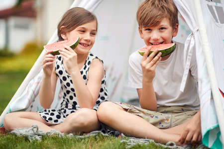 cute boy and girl eating slices of watermelon at picnic in tent. happy childhoodの写真素材