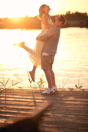 caucasian beardy guy hugging, lifting up his girlfriend at sunset by the riverの写真素材
