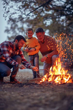 Grandfather, father and grandson enjoying a campfire in the forest on a beautiful autumn duskの写真素材