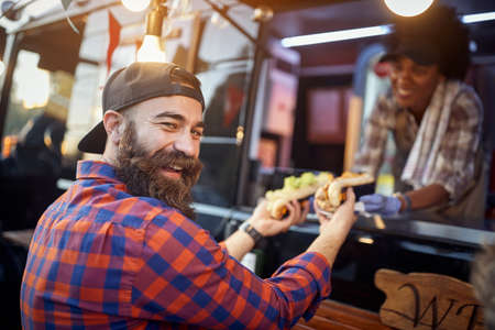 happy, satisfied caucasian beardy male customer taking  sandwiches from a polite employee in fast food service, looking at camera, laughing. eye contactの写真素材