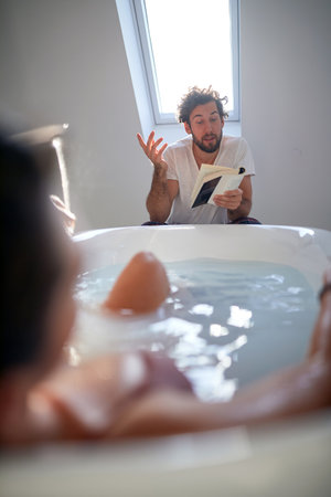 A young guy reading a book to his girlfriend who enjoying the bath in the relaxed atmosphere of the bathroom. Love, relationship, togetherの写真素材