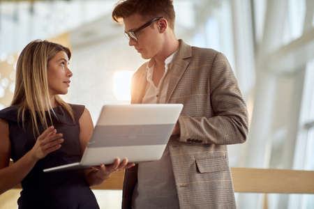 young attractive female holding laptop talking to a male colleague in hallway of business building illuminated with sunlightの写真素材