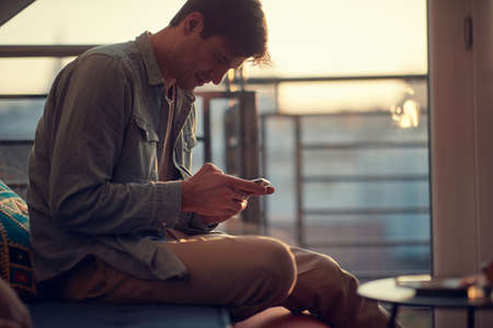 A young guy texting on the smartphone while resting in a pleasant atmosphere at home. Man, home, leisureの写真素材