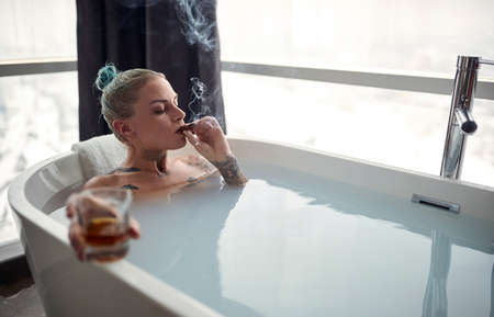 A young sexy tattooed girl enjoying a cigar and a drink while having a bath in a relaxed atmosphere...