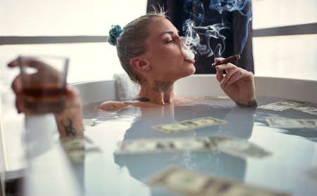 A young sexy tattooed girl enjoying a cigar and a drink while having a bath in a relaxed atmosphere in the bathroom with a bunch of money in the water. Relaxation, luxury, bathroom, homeの写真素材
