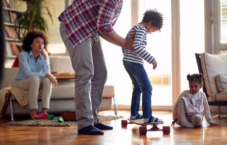 A young father helping his son riding the skateboard in a playful atmosphere at home. Family, together, love, playtimeの写真素材