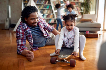 A young father pushing his little daughter on the skateboard in a cheerful atmosphere at home. Family, together, love, playtimeの写真素材