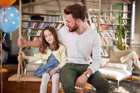 A happy Dad sitting on the swing in a family atmosphere at home with his little daughter. Family, leisure, togetherの写真素材
