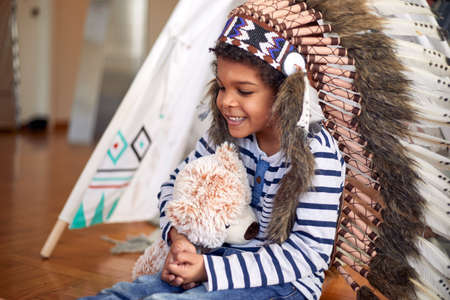 A cute little boy with indian war bonnet holding a teddy while sitting on the floor and playing in a cheerful atmosphere at home. Family, home, playtimeの写真素材