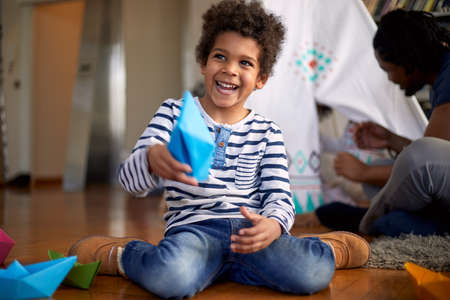 A cute little boy playing with paper boats on the floor and posing for a photo while spending a time with his family in a relaxed atmosphere at home. Family, home, playtimeの写真素材