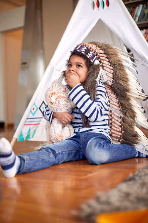 A cute little boy with indian war bonnet sitting on the foor and playing in a cheerful atmosphere at...