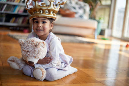 A cute little girl with a crown on her head holding a teddy and posing for a photo while playing in a relaxed atmosphere at home. Family, home, playtimeの写真素材