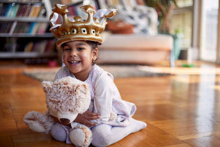 A cute little girl with a crown on her head sitting on the floor in a relaxed atmosphere at home and posing for a photo while hugging a teddy bear. Family, home, playtimeの写真素材