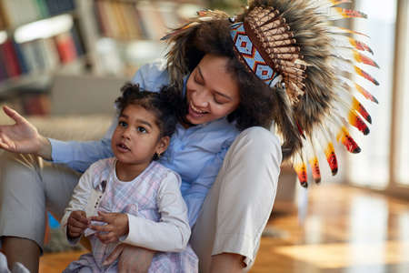 A young mother with indian war bonnet sitting on the floor in a cheerful atmosphere at home and playing with her little daughter. Family, home, playtimeの写真素材
